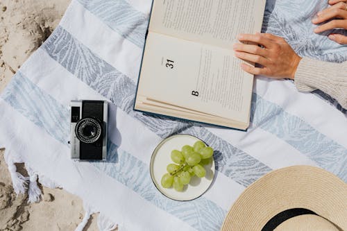 A Person Holding an Open Book Beside an Analog Camera and Green Grapes