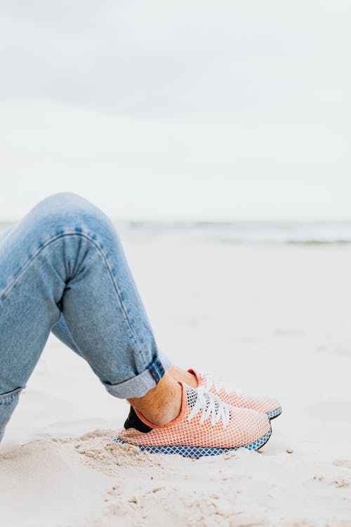 Person in Blue Denim Jeans Wearing Sneaker on the Sandy Shore