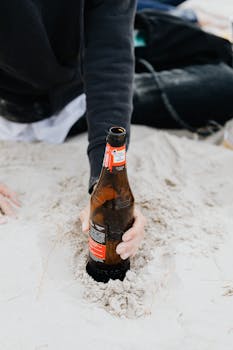 Hand holding a beer bottle placed in the sand, capturing a relaxed beach moment.