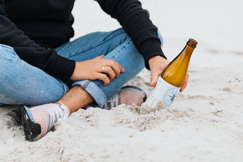 Free Person in Black Jacket Sitting on the Sandy Shore while Holding a Glass Bottle Stock Photo