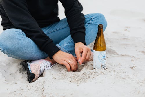 Free Person in Black Jacket and Denim Pants Sitting on a Sandy Shore Beside a Glass Bottle Stock Photo