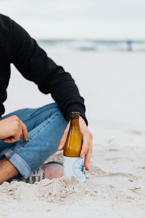 Person in Black Jacket Holding Glass Bottle while Sitting on a Sandy Shore
