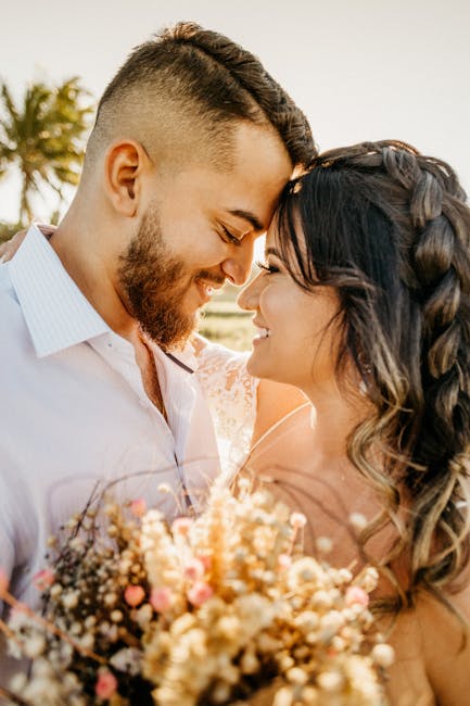 Side view of romantic young ethnic groom and bride touching foreheads and hugging each other while standing on beach with flowers in hand during wedding celebration