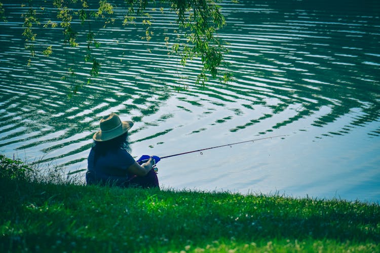 Person Fishing On Green Grassy Embankment Of Rippling River