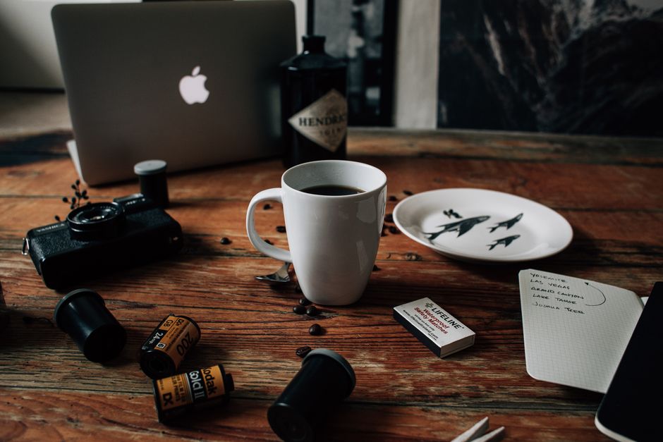 Messy wooden desk with modern laptop old photo camera and films scattered near cup with black fresh coffee