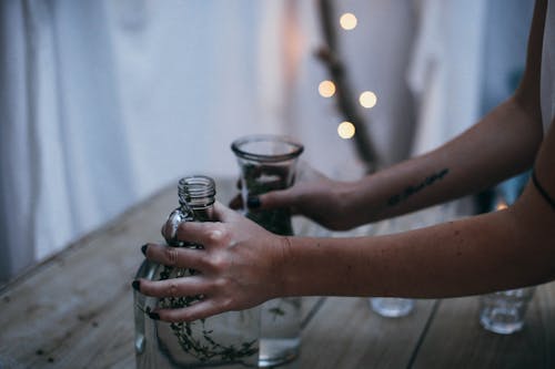 Crop anonymous female putting glass vases on wooden table in creative construction made of hanging white bedsheets and decorated with garlands at twilight