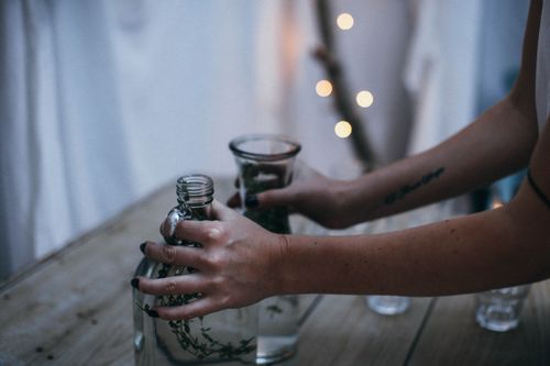 Free Crop anonymous female putting glass vases on wooden table in creative construction made of hanging white bedsheets and decorated with garlands at twilight Stock Photo
