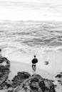 一名男子站在岩石海滩上，在黑白背景下凝视着海浪