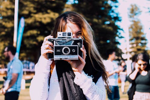 Young female photographer in casual outfit taking pictures on retro photo camera while standing in crowded sunny park