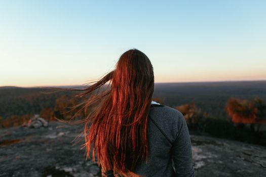 阳光明媚，一位红发、衣着保暖的女旅行者站在山顶，欣赏着下方树木繁茂的山谷