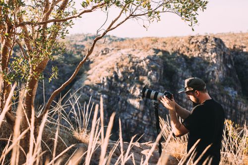 Free Side view of unrecognizable photographer in casual clothes and cap standing on grassy ground near green tree and taking photos of stone hills with photo camera in sunny summer day Stock Photo