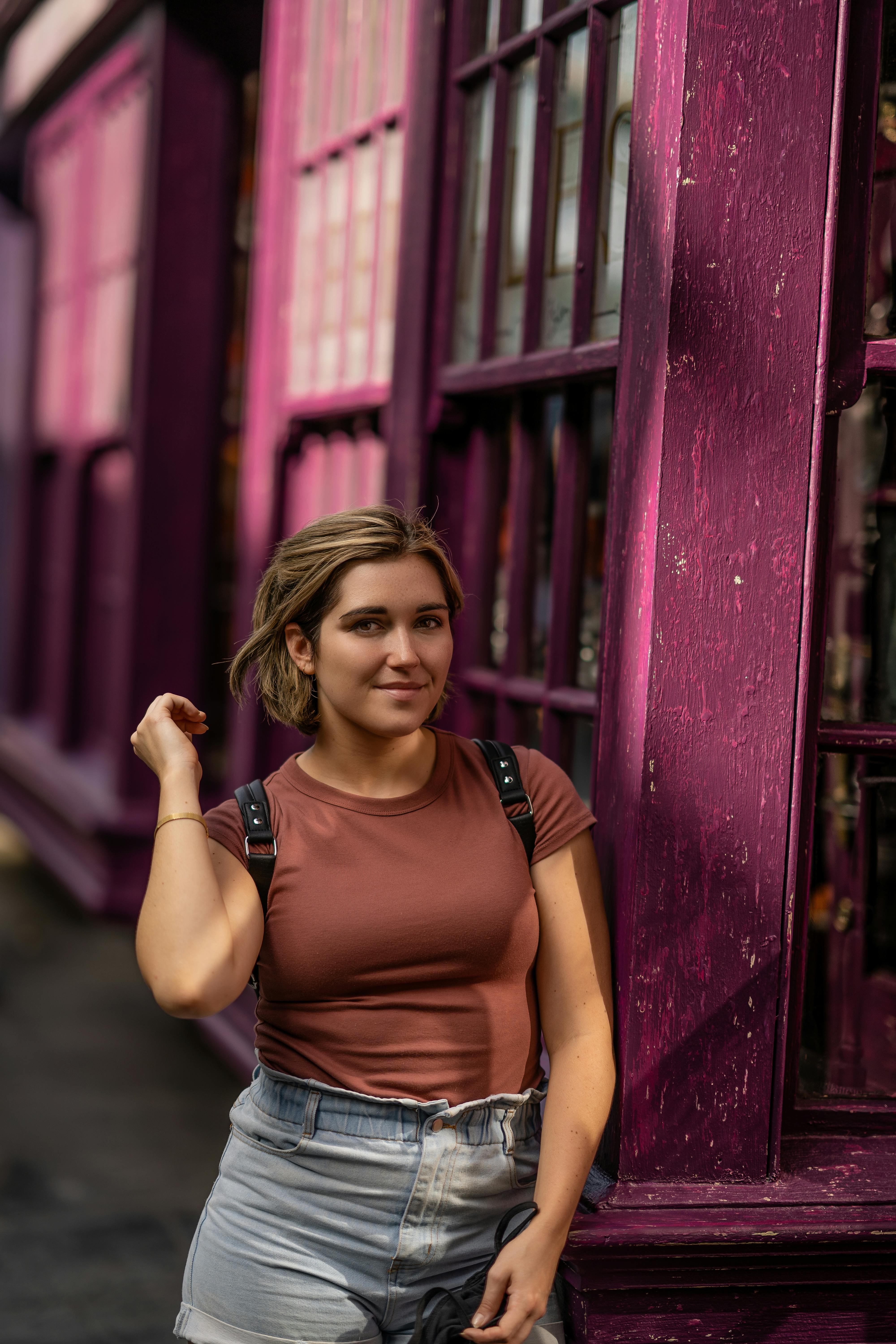 Happy female in casual outfit with hand raised smiling and looking at camera while walking along building with purple wall on street