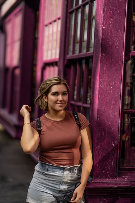 Happy female in casual outfit with hand raised smiling and looking at camera while walking along building with purple wall on street
