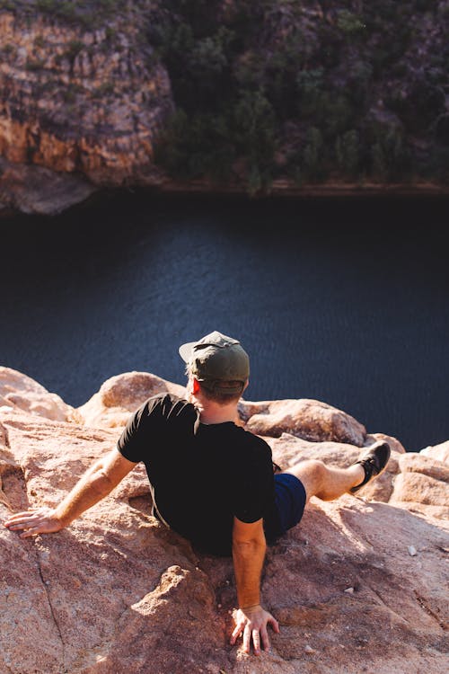 Free Back view faceless male traveler wearing black casual clothes and cap resting on rocky cliff near rippling tranquil river on sunny weather Stock Photo