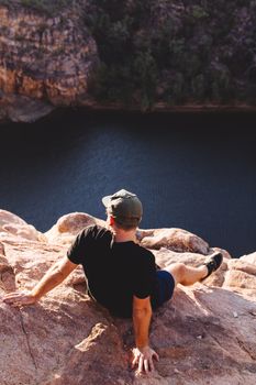 在波光粼粼的宁静河流旁，一位穿着黑色休闲服、戴着帽子的无脸男性旅行者，正背对阳光，在岩石悬崖上休息