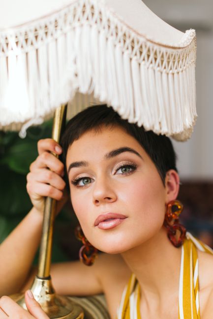 Young alluring female with short hair in earrings looking at camera under lamp with fringes