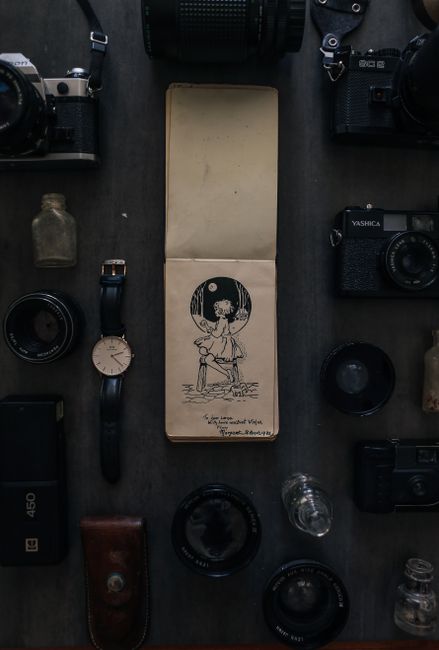 Top view of retro notepad with illustration among professional photo cameras and wristwatch with bottles of perfumes on gray background