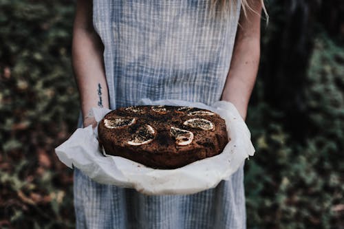 Crop unrecognizable female with tattoo and tasty chocolate cake with lemon slices on top outdoors