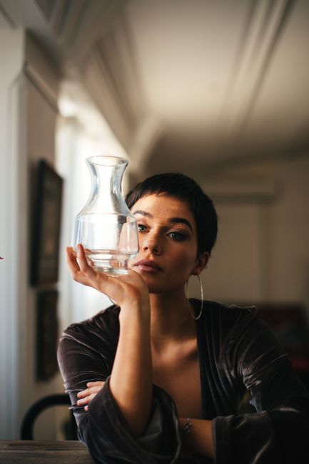 Woman with short hair and hoop earrings contemplatively holding a glass pitcher in a stylish indoor setting.
