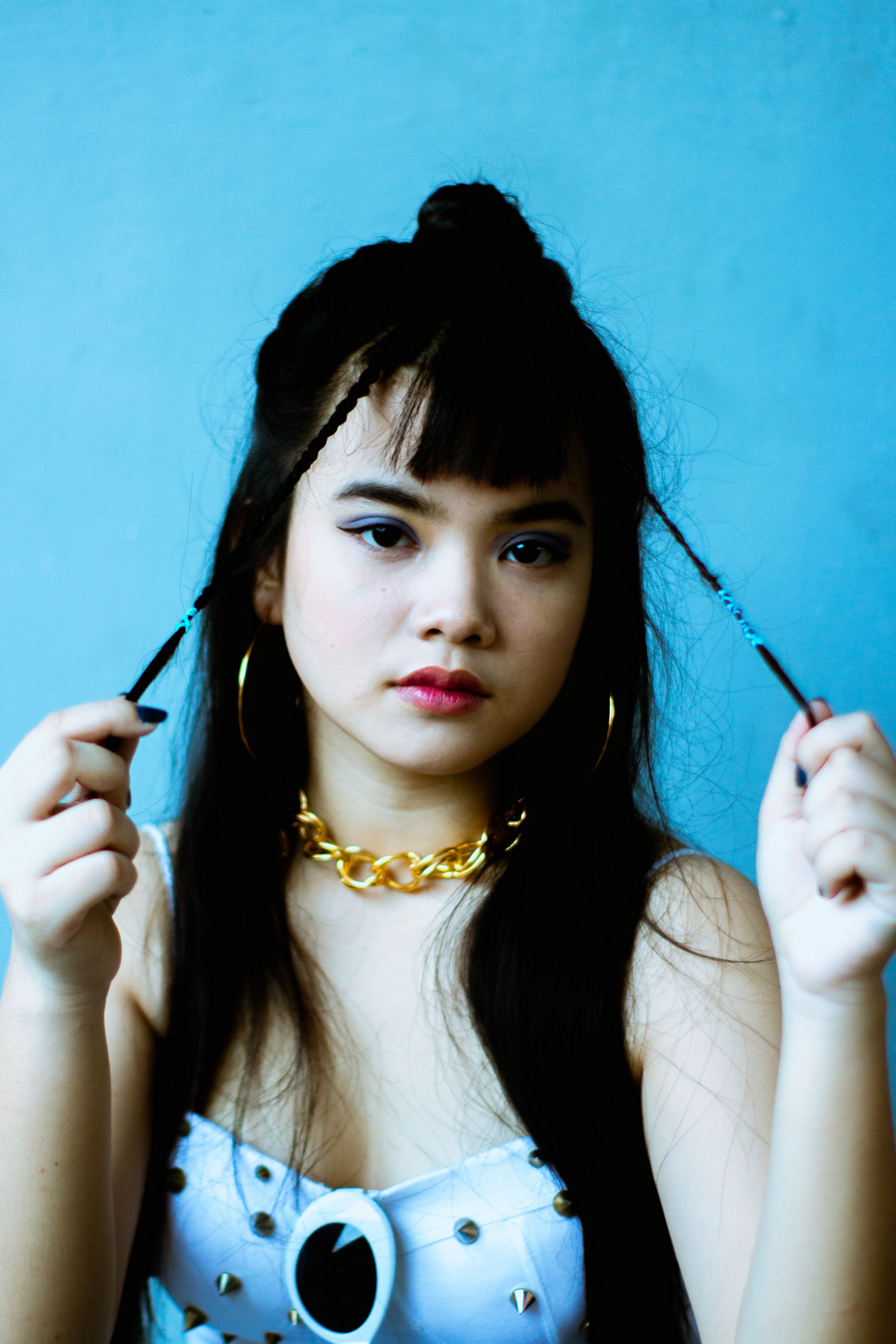 Portrait of a young woman in fashionable attire with braided hair and jewelry against a blue background.