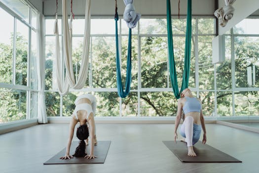 Two women doing yoga poses on mats in a sunlit studio with hanging hammocks.