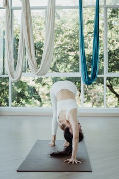 Woman in activewear stretching on a yoga mat indoors with aerial hammocks in the background.