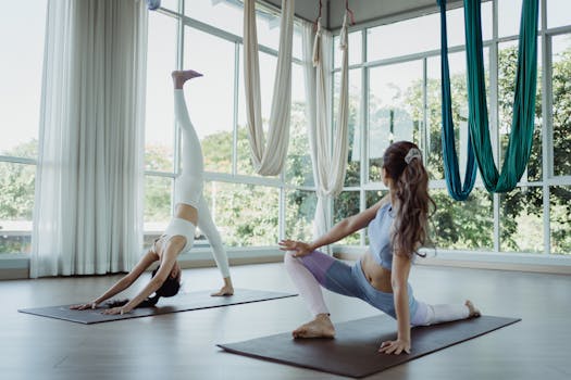 Two women practicing yoga poses in a bright, airy studio with aerial silks and large windows.