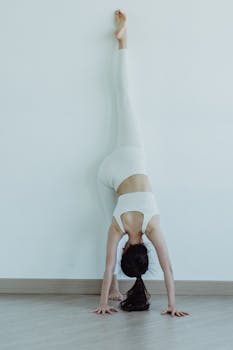 A woman practicing a yoga handstand against a white wall indoors, showcasing strength and balance.
