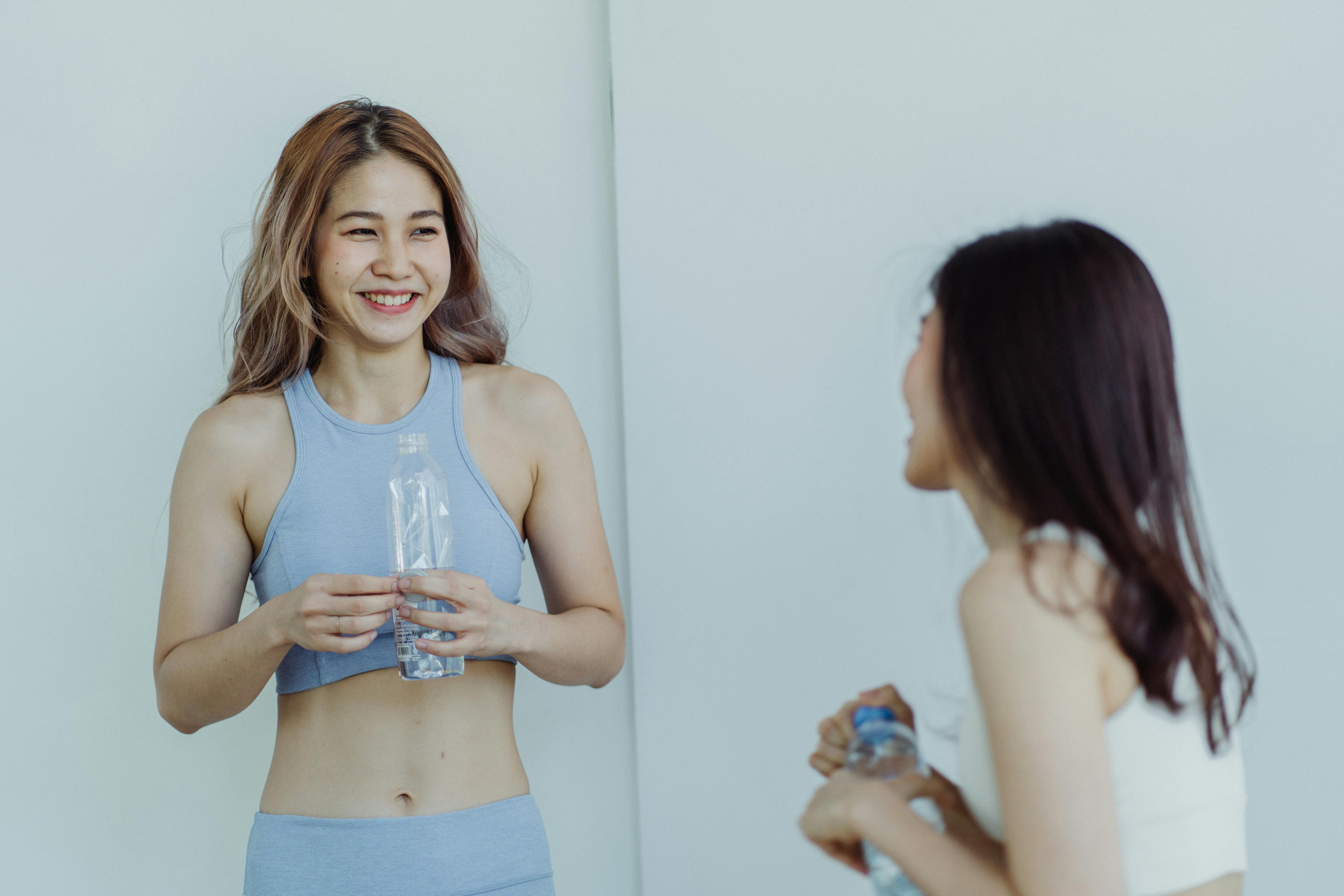 Two women in sportswear smiling and talking indoors, holding water bottles after a workout.