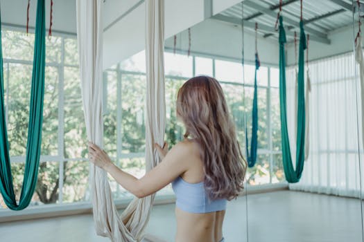 Young woman in sports bra practices aerial yoga with silk hammocks in a bright studio.
