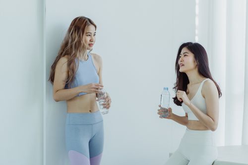 Free Two women in sportswear holding water bottles and chatting in a bright indoor setting. Stock Photo