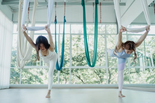 Two women engaged in aerial yoga, stretching using fabric in a sunlit studio.