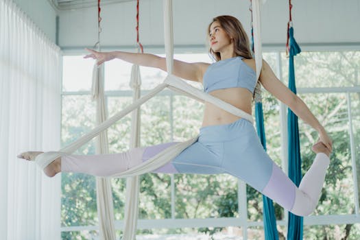 Asian woman performing aerial yoga pose in a bright fitness studio.