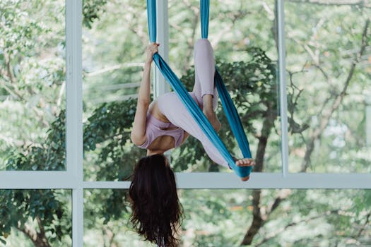 Woman practicing aerial yoga using silk hammock by large window indoors.