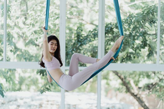 Asian woman performing aerial yoga on a hammock inside a bright room with glass windows.