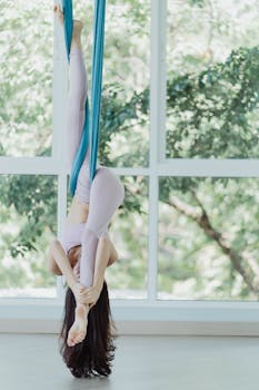 A woman performs aerial yoga suspended upside down in a bright studio.