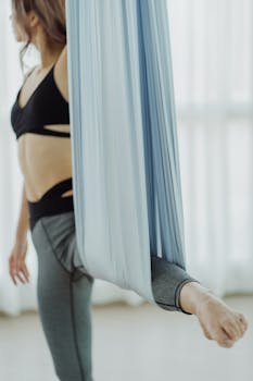 A woman practicing aerial yoga using a silk hammock indoors, focusing on leg stretching and balance.