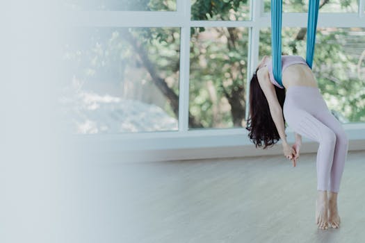 Woman practicing aerial yoga with blue hammock in a sunlit studio, showcasing flexibility and grace.