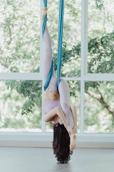 Woman practicing aerial yoga with blue silk in a serene, sunlit studio setting.