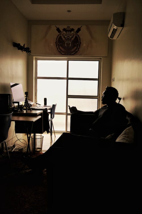 Silhouette of unrecognizable person resting on couch in front of computer under air conditioner in dark room