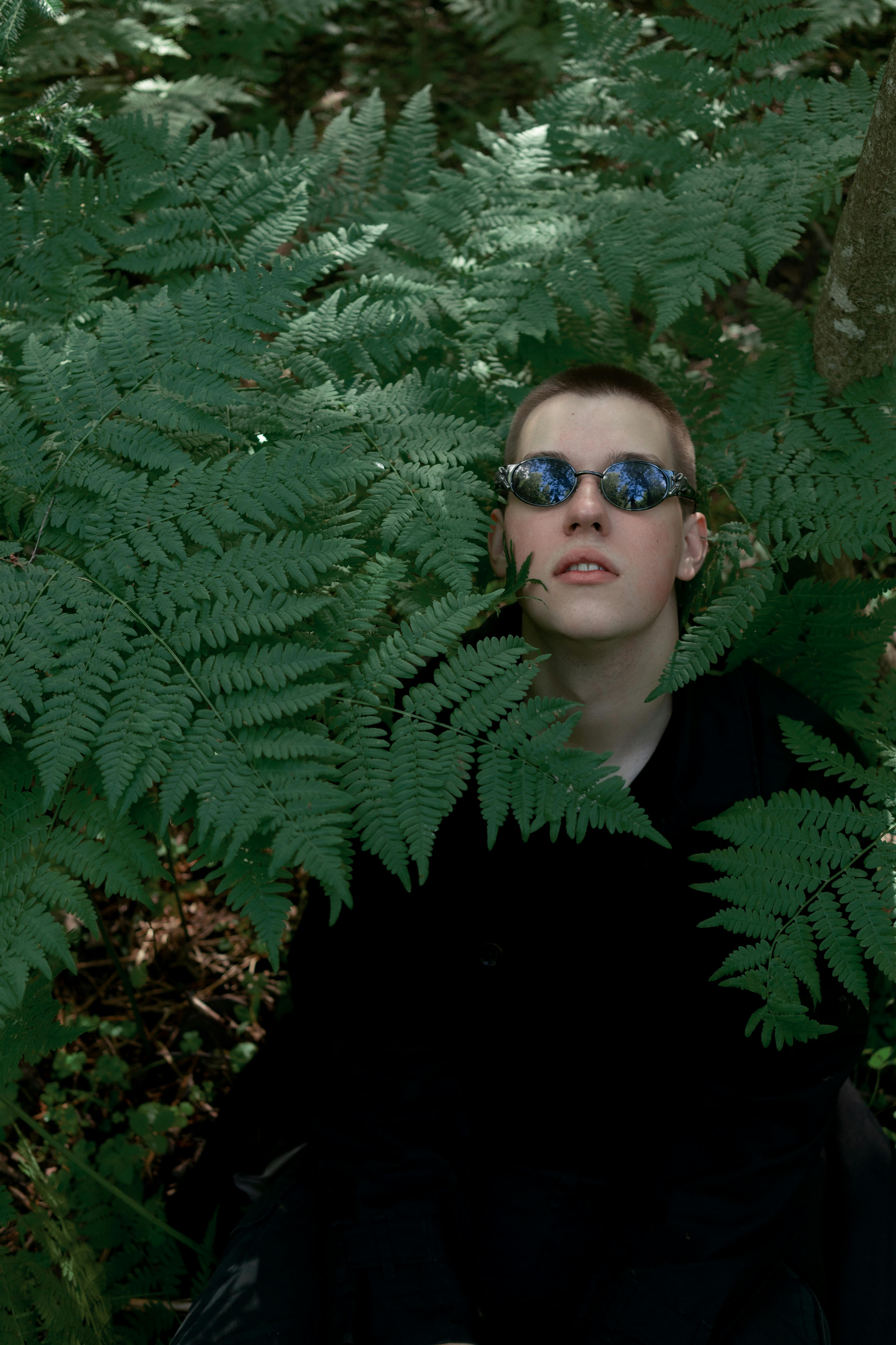 Young trendy male in black wear and sunglasses standing among lush green plant leaves in forest