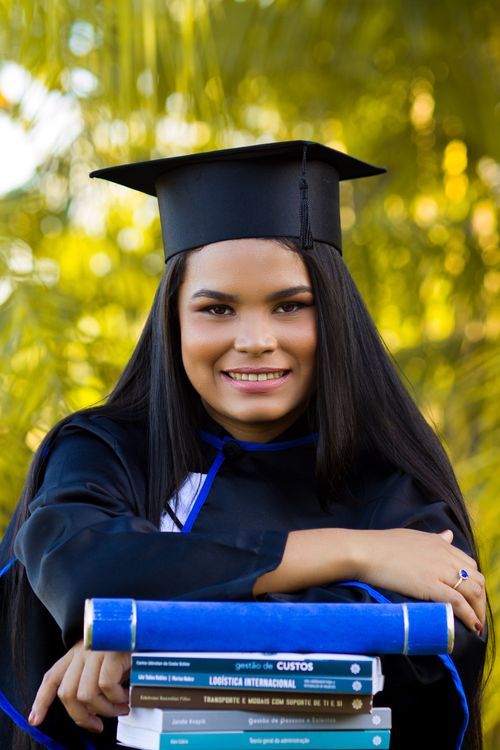 Free Glad young ethnic woman in graduation cap with textbooks smiling and looking at camera on blurred background of greenery Stock Photo