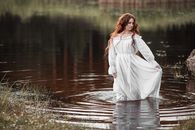 Gorgeous female with long ginger hair and in white elegant dress standing in water and looking down in daylight