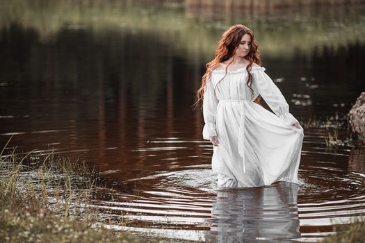 Gorgeous female with long ginger hair and in white elegant dress standing in water and looking down in daylight
