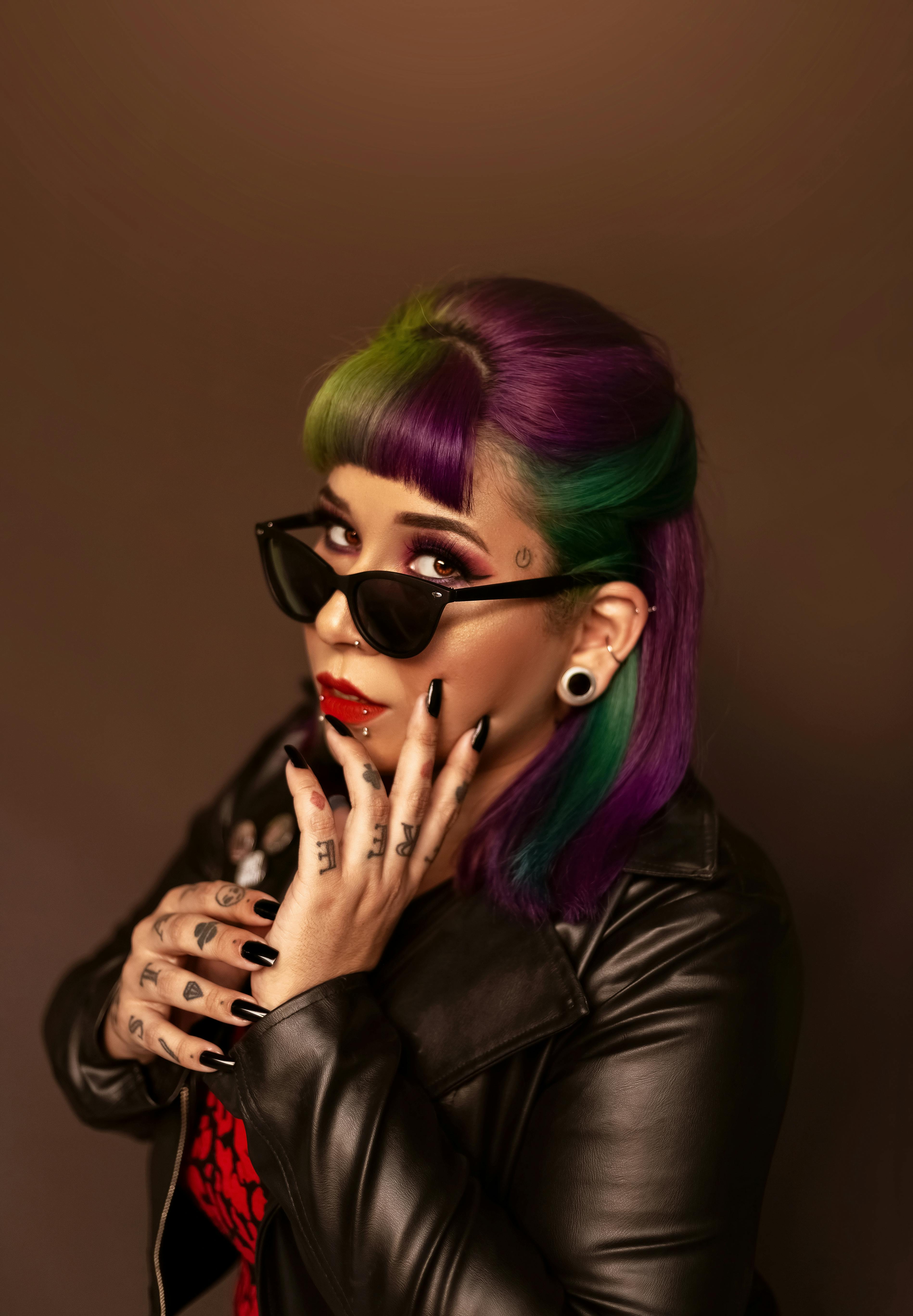 Confident woman with multicolored hairstyle and tattoos  in trendy sunglasses holding hand on cheek and looking at camera