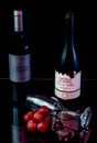 Two red wine bottles with grapes and a glass on a reflective surface.