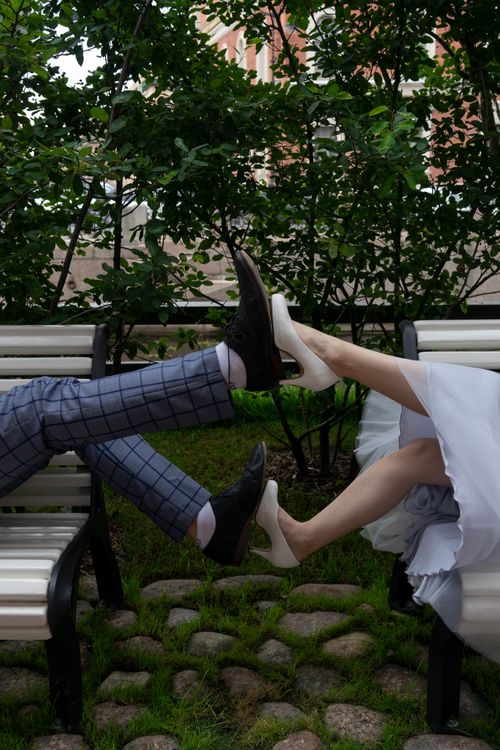 Free Side view of crop unrecognizable stylish groom and wife touching legs while lying on street benches on wedding day Stock Photo