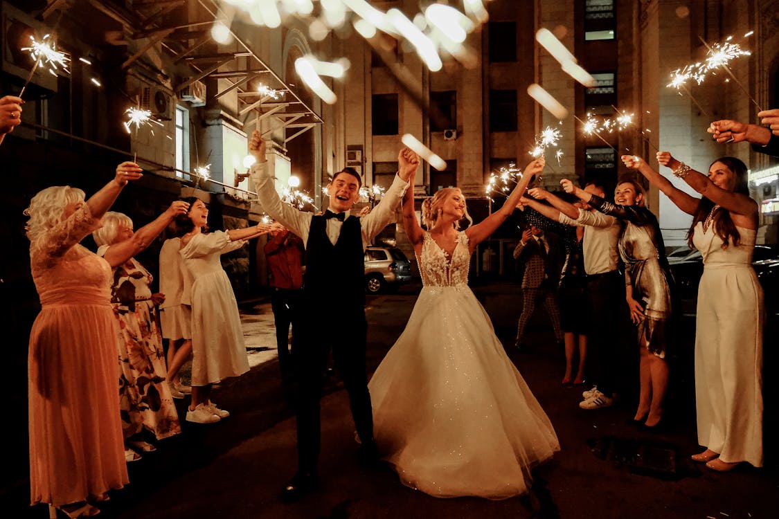 Free Cheerful young bride and groom with guests dancing on street with sparklers in hands during wedding party Stock Photo