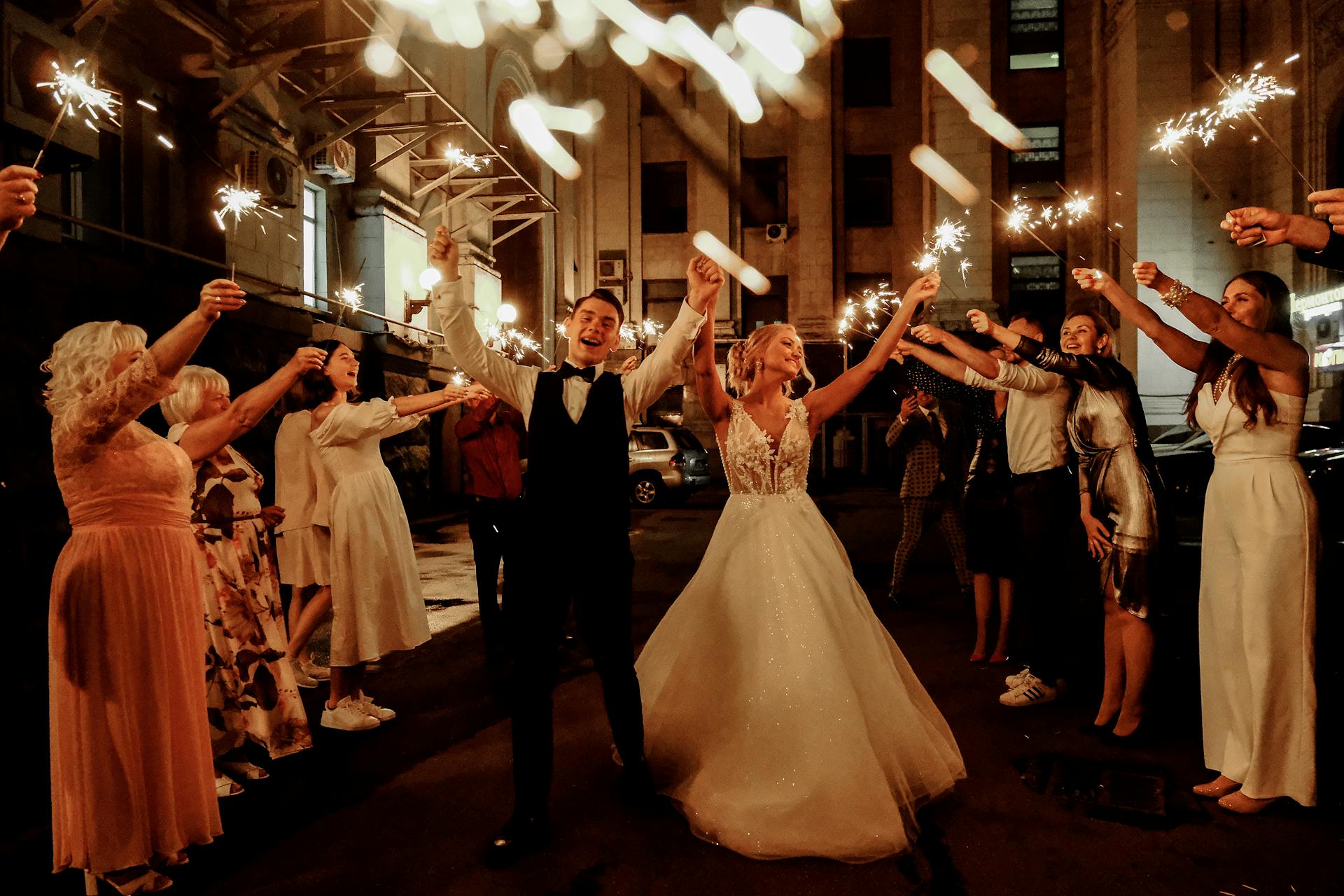 Cheerful young bride and groom with guests dancing on street with sparklers in hands during wedding party