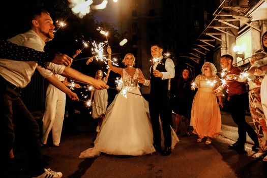 Full body of joyful young bride and groom with sparklers in hands walking near happy guests during wedding celebration in evening
